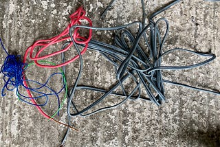 A variety of different insulated electrical wires on a concrete floor. Red, green, blue, and grey. The insulation is stripped of the end of each one.