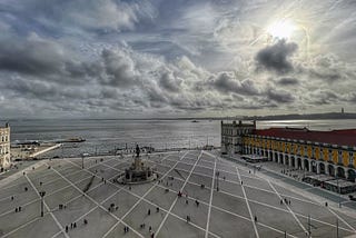 A wide-angle view of a large public square in Lisbon, Portugal, called Praça do Comércio. The square is paved with geometric patterns and has a central statue surrounded by people walking. On the right, there are buildings with yellow facades and a red roof, and in the background, a body of water stretches towards the horizon under a dramatic cloudy sky, with the sun peeking through.