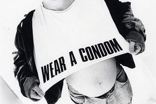 Black and white photo of Sinead O’Connor in a jacket and jeans, wearing a white shirt with WEAR A CONDOM printed on it. By Kate Garner in 1986.