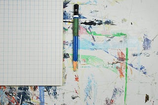 A blank grid notebook laying open on a paint-splattered desk with a pencil next to it.