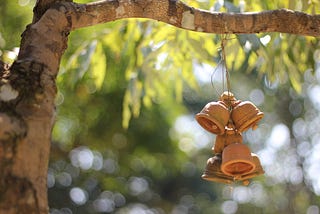 An image of a tree branch in the sunlight. From the branch hang three small, light-brown ceramic prayer bells. The bells hang from a thin string in a bunch, catching the sunlight along their tops.
