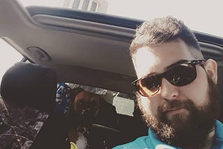 A very handsome man in the foreground (in a truck), holding coffee, wearing sun glasses while his cute Boxer dog looks at the camera as well from the back seat.