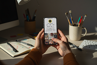 Hands holding a phone showing the Ladies Who Design TikTok profile, with art supplies and a sketchbook on the desk in soft afternoon light