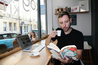 A casually dressed man in his late-twenties sitting in a cafe. He has a laptop open on his table, and he is sipping an espresso while reading a book called, “How the World Thinks.”