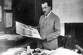 A man, Nikolai Vavilov, stands over a desk in a suit, holding up a scientific print. His desk is covered in plant samples