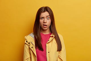 portrait of a disappointed and shocked woman. One eyebrow is raised and she is smirking as if she has seen something unpleasant. She has long brown hair and is wearing a red jumper under a yellow coat, against a yellow background