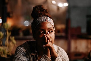 A Black person sitting a table, with their hands laced together in front of their face. They’re wearing a patterned headwrap, and their braids are tied up in a bun high on their head. They’re looking to their left. They’re wearing an off-white shirt under a beige knit sweater.