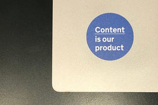 A blue sticker that says ‘Content is our product’ on a silver laptop