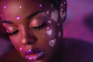 A Black woman, eyes closed with a short afro, with pink rhinestones on her face, white hearts pressed on her cheeks and sparkly pink lipstick. Yummy.