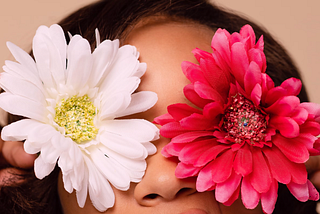 A black woman holds two flowers over her eyes. The right one is a white flower and the left one is a pink flower.