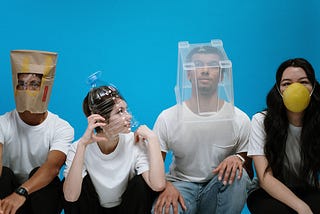 four people sitting with various face coverings (plastic bottle, mask, etc)