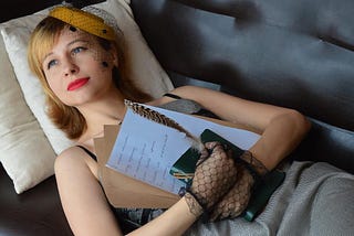 Photo of a young woman lying on a sofa and holding a journal, a feather pen, and a book to her chest as she gazes off into the distance with a dreamy look.