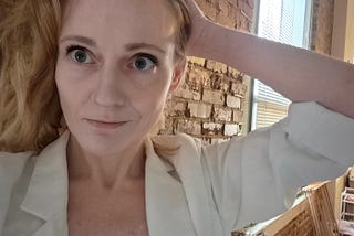 Photo of a blonde woman in a cream-colored blazer standing against a brick wall, holding back the hair from one side of her face with her left hand and slightly smiling.