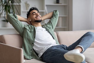 A young Arab man leaning back on a sofa, airpods in his ears, with his hands behind his head and a smile on his face. He is clearly enjoying spending this time by himself.