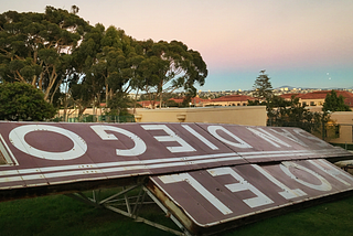 A vintage sign for Hotel San Diego lays on the ground upside down. The city and trees are in the background.