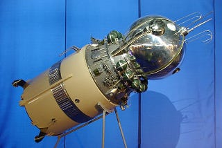Color photograph of a model of the Vostok 1 spacecraft set against a blue background