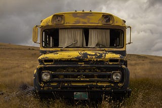 Old, rusted bus, with curtains hanging in the front windows, sitting in a field.