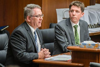 Two men in suits sit behind a wooden table. One of them speaks into a microphone as the other looks on.