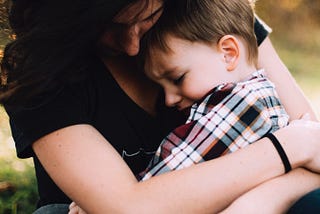 Mother holding a young child on her lap and hugging him close in a reassuring manner. The mother and child’s face are cast down. The mother is wearing a dark shirt and the son is wearing a plaid patterned shirt.