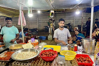 Two cooks at a food cart work —  one who is slicing onions against a well-worn piece of wood, and another continues to cook the red chicken nuggets from the earlier picture