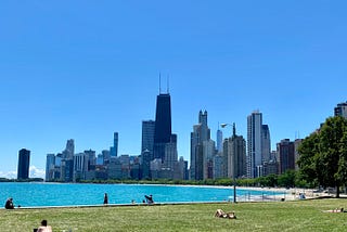 Image of the Chicago skyline and Lake Michigan on a bright, warm summer day. A few people are lounging in the park that borders the lake.