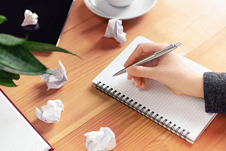 A person with a pen poised to write on blank paper in a notebook with 4 crumpled up sheets of paper next to it.