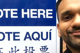 Anu is standing next to a Vote Here sign and smiling