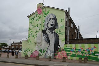 Large mural on a wall of the side of a building on an housing estate depicting a portrait of guitarist Mick Ronson, head and upper body, surrounded by flowers and greenery, also showing children on a park’s running track.