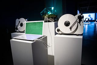 A laptop, two vinyl players and the moss installation each of them placed on a white pillar in an exhibition space.