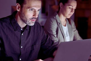 Zdjęcie przedstawia osoby pracujące przy laptopach w nocy. Mężczyzna ubrany w ciemną koszulę, ma skupiony wyraz twarzy i patrzy intensywnie w ekran, co sugeruje pełne zaangażowanie w wykonywane zadanie. W tle widać kobietę w jasnej marynarce, również pochyloną nad laptopem, co wskazuje na jej koncentrację i determinację. Obraz oddaje istotę wyzwania, jakim jest praca w nocy osób niepełnosprawnych — skupienie, determinację oraz wytrwałość w dążeniu do celu pomimo zmęczenia.