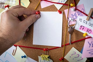 A hand pins a notecard onto a bulletin board covered in sticky notes and red string.