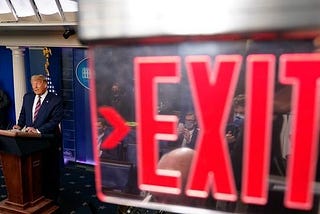 Photograph of Trump at the podium in the white house press room in the background of an Exit sign.