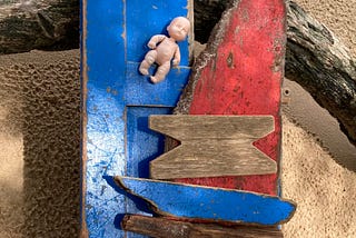 Foto colorida, ext, dia. Uma tábua velha azul e uma vermelha estão apoiadas sobre um tronco de árvore sobre areia. Nas tábuas, há pequenas lascas de madeiras horizontais e paralelas. Na parte superior da tábua azul, há uma pequena miniatura de boneca de plástico.