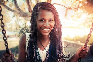 Photo of a smiling young woman with flowing braids. She is on a swing with the sun shining behind her.