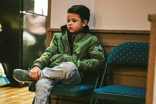 A young boy sits alone on a folding chair with his arms crossed and legs tucked in, wearing a green jacket and jeans. His expression looks distant and serious, as though he’s deep in thought or waiting.