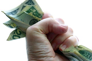 Photo of a hand, against a solid white background, tightly clutching a number of bills of different U.S. denominations.