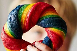 A white hand holds up a rainbow-coloured bagel to the camera, their face in the background is blurry