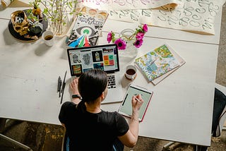 A woman sits at a table with her computer and sketch pad creating art.