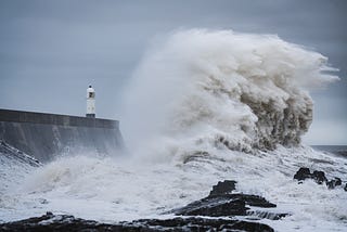 An enormous wave crashing against the shore, where a small lighthouse stands.