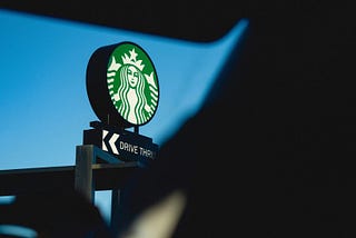 Image shows iconic green and white Starbucks sign reading “Drive Thru” against a bright blue sky background, viewed through the front windshield of a car. Foreground shows blurred, dark silhouette of car’s interior, including seat, visor, and steering wheel.