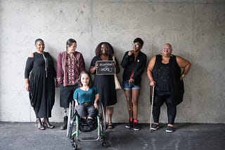 Six disabled people of colour smile and pose in front of a concrete wall.