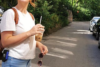 Christina smiler bredt til kameraet, med en iskaffe i den ene hånden, og solbrillene i den andre. Hun har en sekk på ryggen og en lysmåler hengende over skuldrene.