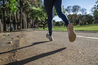 Legs and feet of an athlete running down a road in a green park surrounded by trees.