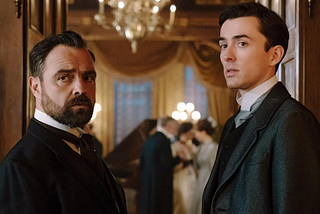 The lead characters of Vienna Blood; two white men in Edwardian suits in a fine room with chandelier and piano.