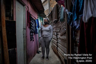 Brazilian woman stands in an alleyway, wearing a cotton mask, a grey top and pants. Colorful laundry hangs on either side.