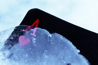 The left top corner of an Ace of Hearts card rises out of the ice. The card is black with a red A above a red heart. The ice is pale blue.