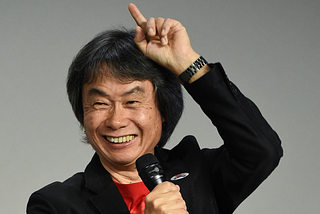 Shigeru Miyamoto on stage, pointing to the sky with a microphone in his hand.
