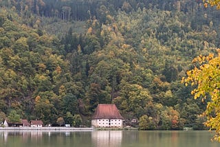 A building with red roof on the river bank, with autumn trees above.