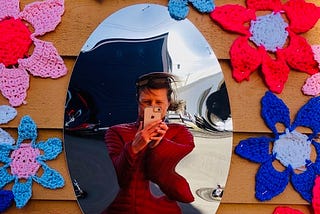 Nina Elliott takes a photo of herself in a distorted mirror surrounded by colourful crocheted flowers hung on an outside wall.