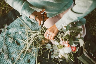 A cropped image of a woman in a soft turquoise sweater cutting flower stems in her lap.